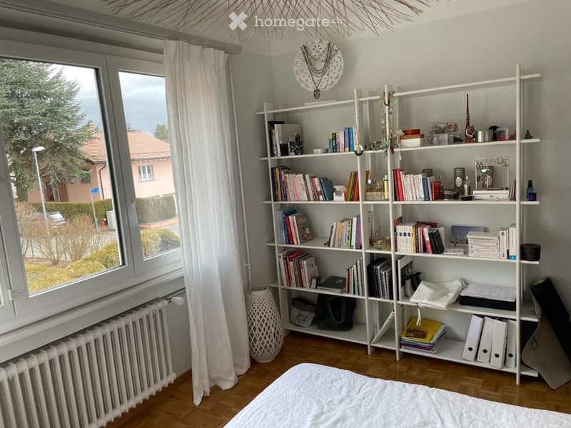 A well-lit bedroom with a wooden floor, a large window with white curtains, a white bookshelf filled with books and decorative items, a white bed, and a decorative vase near the window.