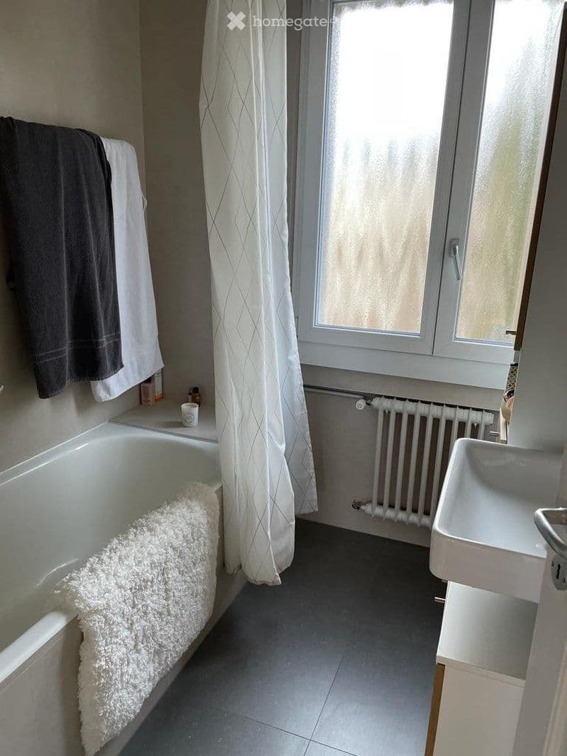 Bathroom with a bathtub, sink, radiator, and a window with a curtain. Towels are placed on the edge of the bathtub and on a rack.