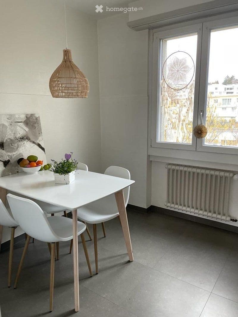 Modern dining area with a white dining table, four white chairs, a bowl of fruit, a small potted plant, and a hanging woven light fixture.