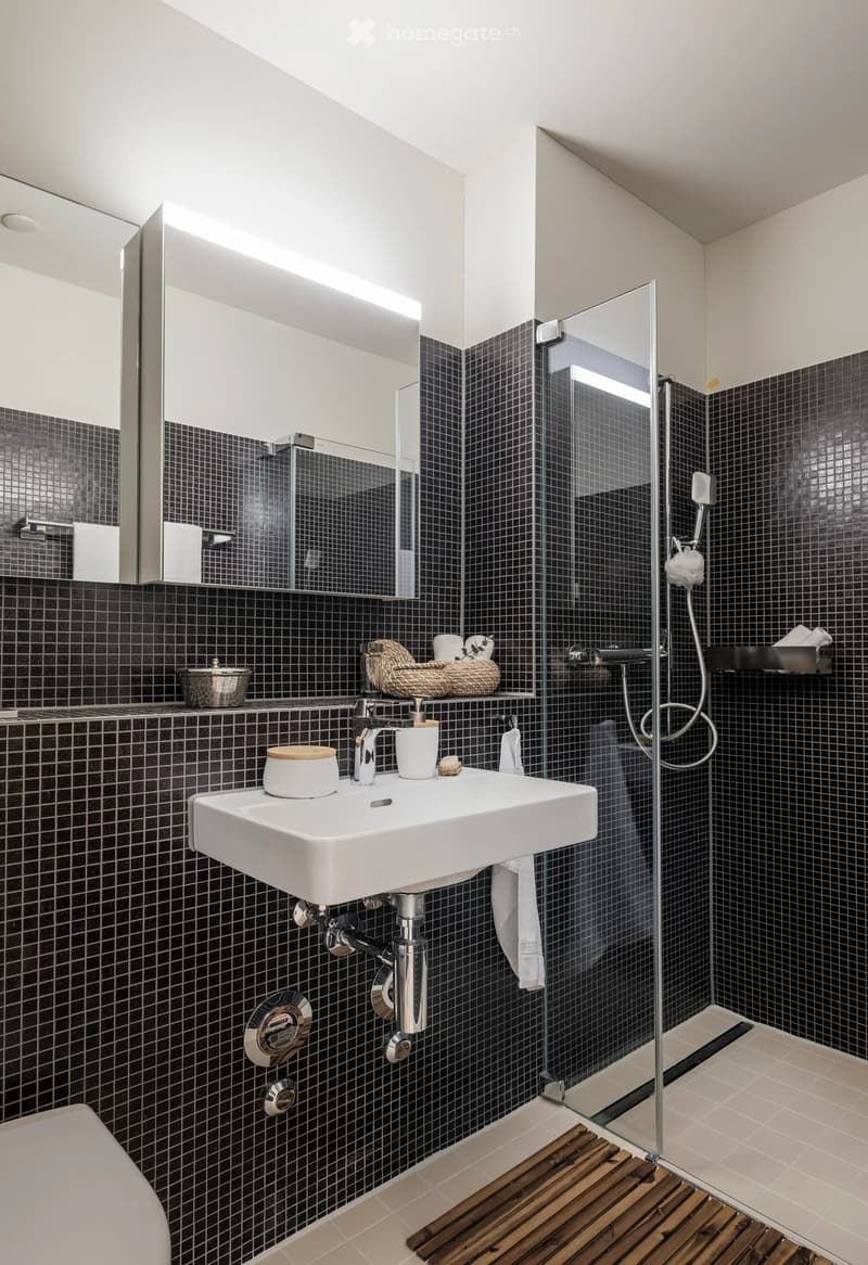 Modern bathroom with black tiled walls, white sink, glass shower enclosure, and wooden bath mat.
