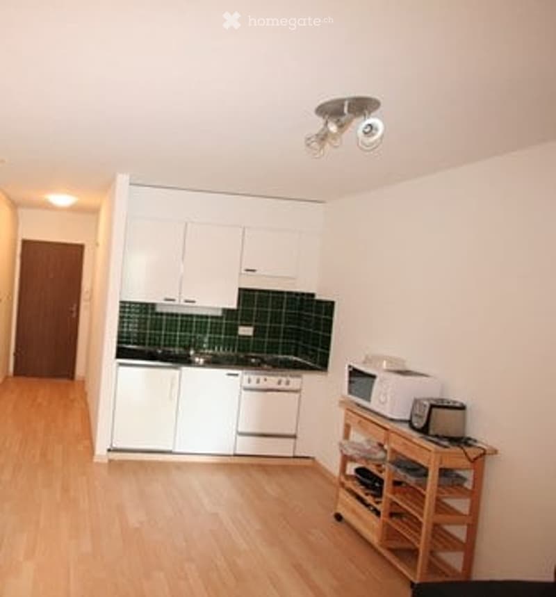 Modern kitchen with white cabinets, green tiled backsplash, wooden flooring, and small wooden shelf with appliances.