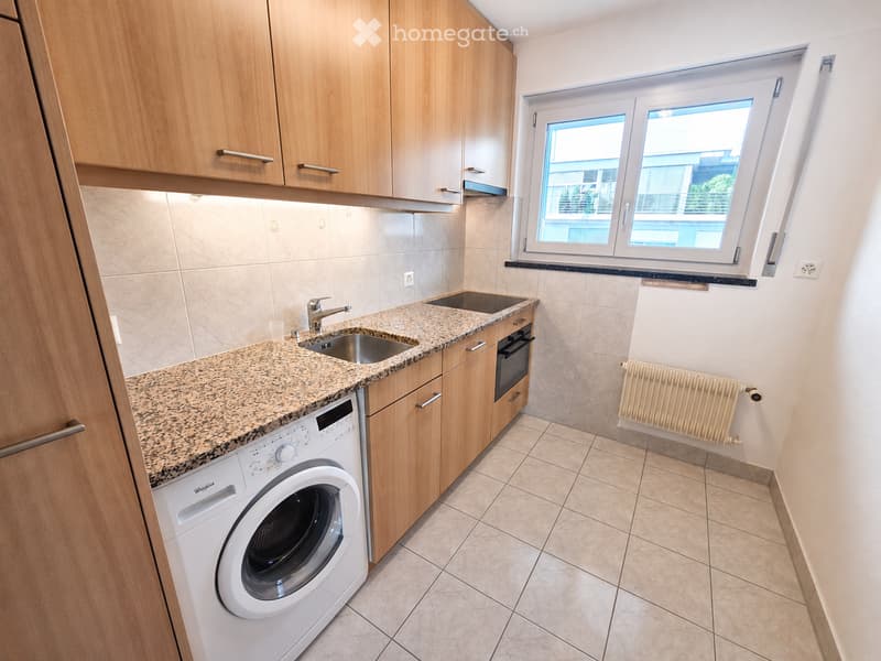 Modern kitchen with granite countertop, stainless steel sink, wooden cabinets, built-in oven, and washing machine. Features a window with a view of the outside.