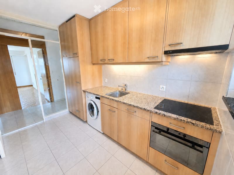 Modern kitchen with light wooden cabinets, granite countertop, stainless steel sink, induction cooktop, built-in oven, and washing machine. The space is bright with tiled flooring and a visible hallway beyond a glass door.