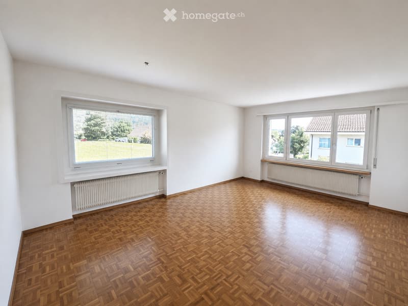 Empty room with wooden herringbone flooring, white walls, large windows with blinds, and radiators beneath the windows.