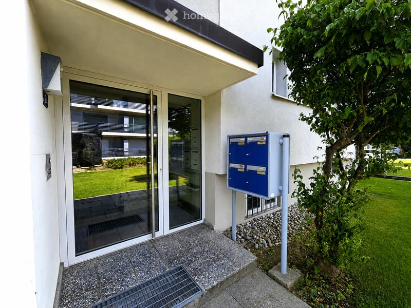 Entrance to a modern building with a glass door, mailboxes, and a small garden area with a tree.
