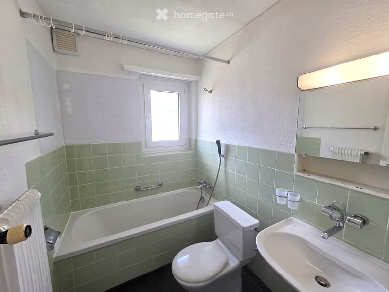 Bathroom with green tiled walls, bathtub, toilet, sink, mirror, and two cups on the wall.