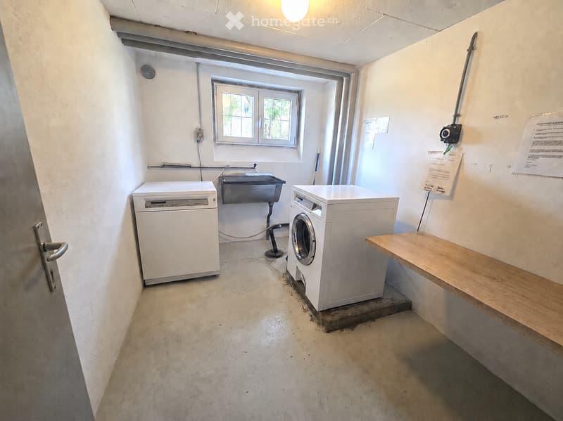 Utility room with a washing machine, dishwasher, sink, and a wooden bench. The room has concrete flooring, white walls, and a window with a view of greenery.