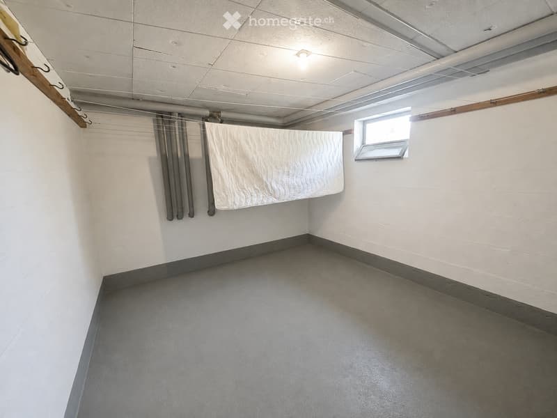 Empty room with white walls, grey floor, and a small window. A single mattress is leaning against the wall.