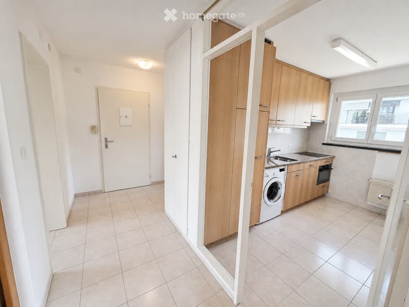 The image shows a modern, unfurnished kitchen with light wooden cabinets, a sink, an oven, and a washing machine. The space features tiled flooring and white walls, with a door leading to another room.