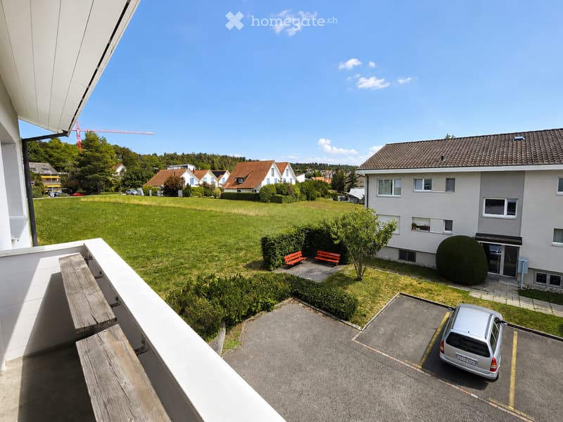 Balcony overlooking a green lawn, with a parking area below and a silver car parked. The surrounding area includes residential buildings and a clear blue sky.