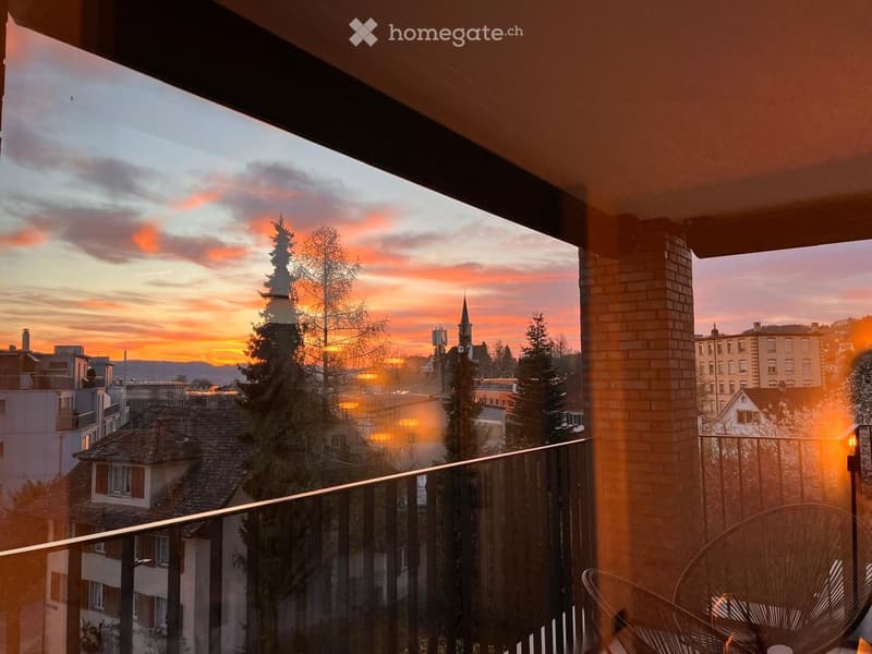 Balcony with a view of a cityscape at sunset, featuring trees, rooftops, and a distant church spire.