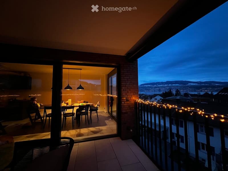 Balcony with a view of the city and mountains, decorated with string lights, connected to a dining area with a table and chairs.