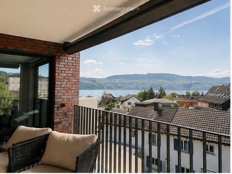 Balcony with a scenic view of a lake and surrounding mountains, featuring a wicker chair with cushions and a metal railing.