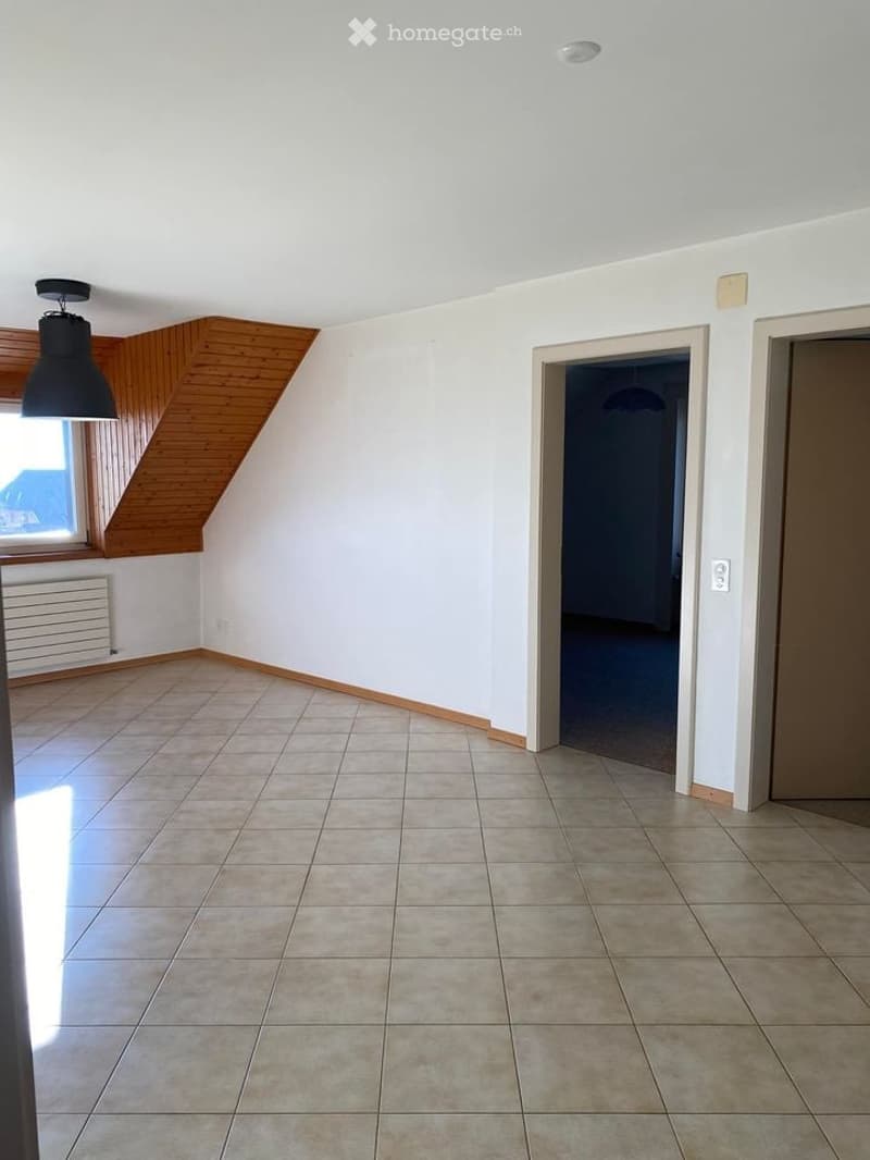 Empty room with tiled floor, white walls, and wooden slanted ceiling. Features include a window with wooden frame, radiator, and two doors leading to other spaces.