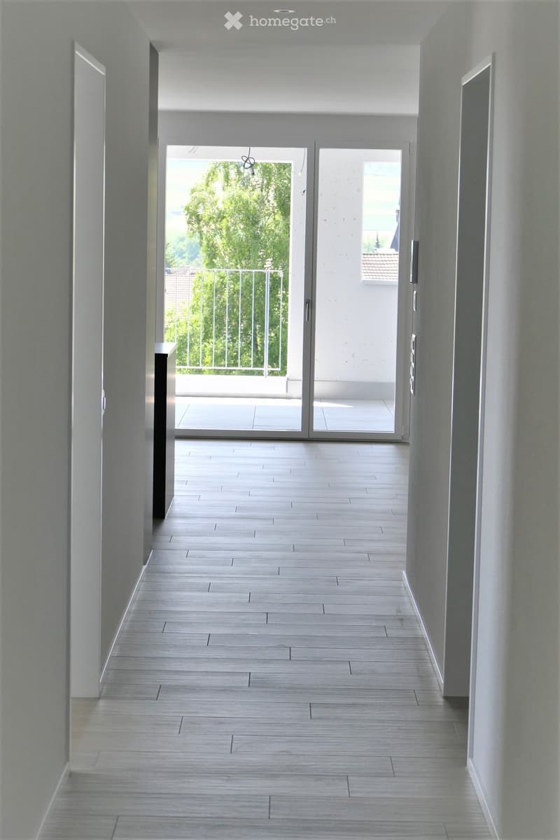 Modern hallway with light-colored tiled flooring, white walls, and a large sliding glass door leading to a balcony with a metal railing.