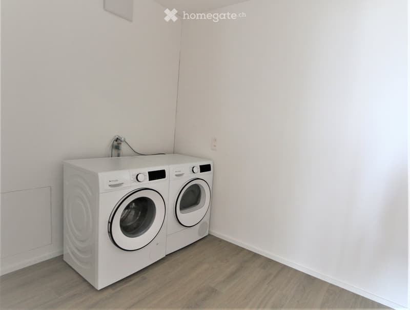 A small, unfurnished room with light-colored walls and wooden flooring, featuring a white washing machine and dryer unit against one wall.