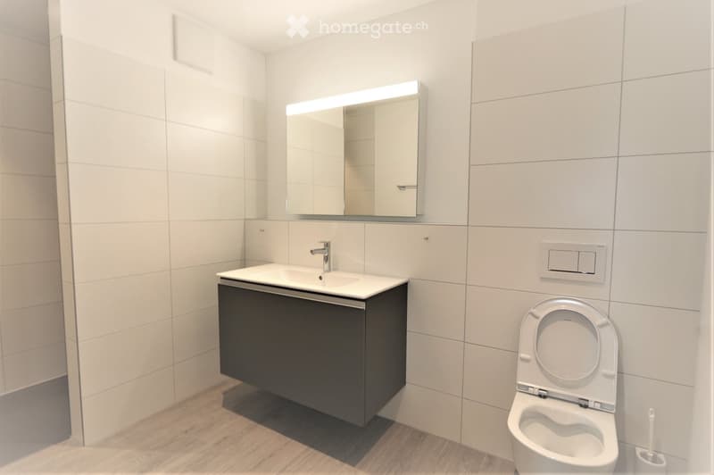 Modern bathroom with white tiled walls, a dark grey cabinet under a white sink, a chrome faucet, a large mirror with built-in lighting, and a toilet with the lid up.