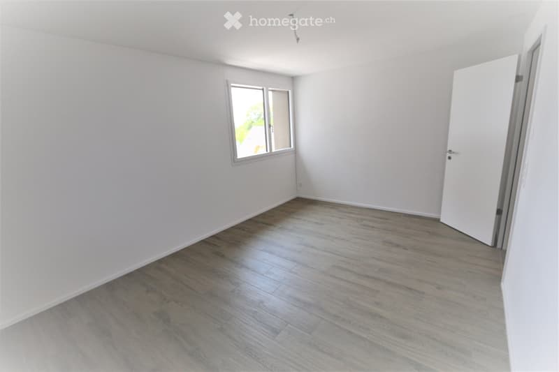 Empty room with light grey walls, wooden flooring, and a single window letting in natural light. The room has a white door on the right side.
