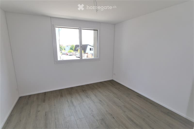 Empty room with white walls, wooden flooring, and a large window with a view of the street.
