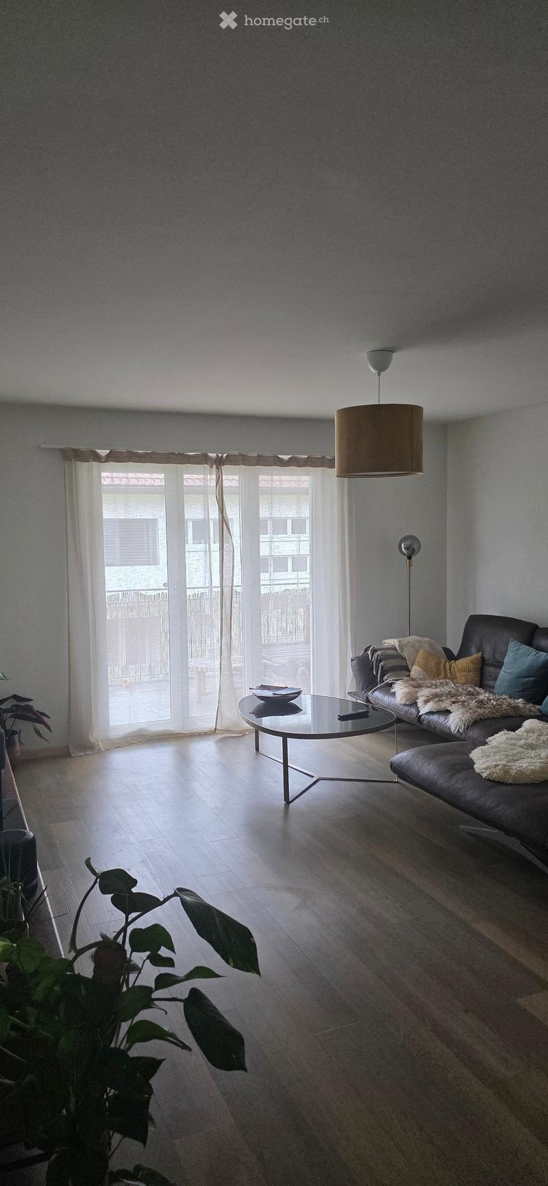 Modern living room with wooden flooring, large windows, a black leather sofa, a glass coffee table, and a floor lamp.