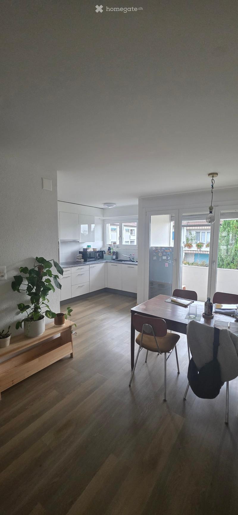 A modern kitchen and dining area with white cabinets, a wooden dining table, chairs, and a potted plant on a sideboard. The kitchen features a sink, microwave, and refrigerator, with large windows and a sliding glass door leading to a balcony.