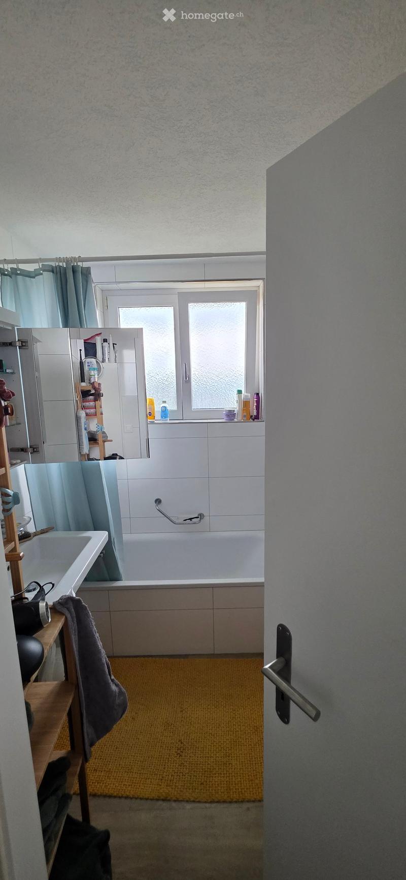 Bathroom with a bathtub, sink, mirror, and shower curtain. Shelves hold toiletries and a towel is draped over a wooden stand.