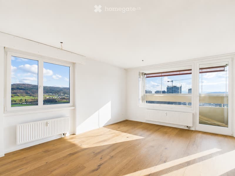 Empty room with wooden flooring, white walls, large windows offering views of both countryside and cityscape, radiators along the walls.