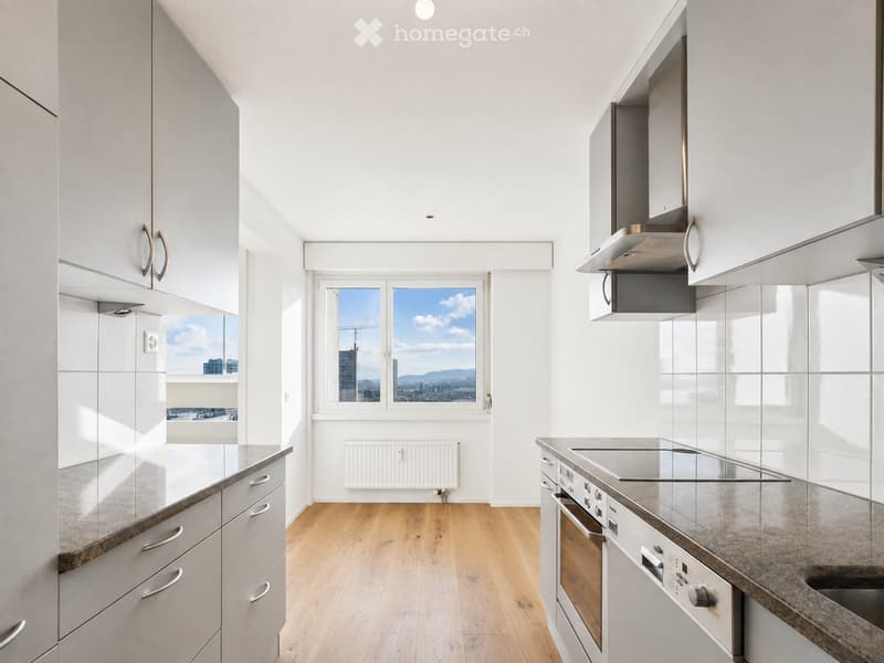 Modern kitchen with white cabinets, stainless steel appliances, wooden flooring, and large windows offering a city view.