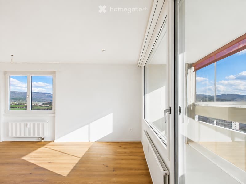 Bright, empty room with wooden flooring, white walls, large windows offering scenic views, and radiators along the walls.