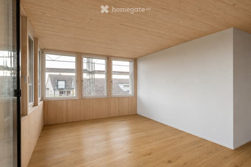Empty room with wooden flooring, white walls, and large windows offering a view of the street and neighboring houses.