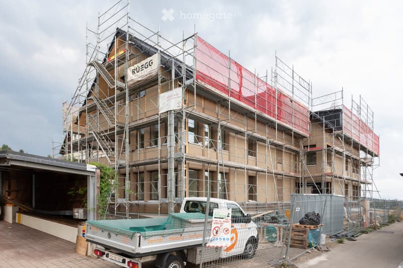 A multi-story building under construction, covered in scaffolding with visible wooden framing and signage for "RUEGG" and "NARROW AG".