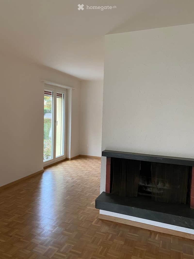 Empty room with wooden herringbone flooring, white walls, and a fireplace with a black surround. A large sliding glass door leads to a garden area.