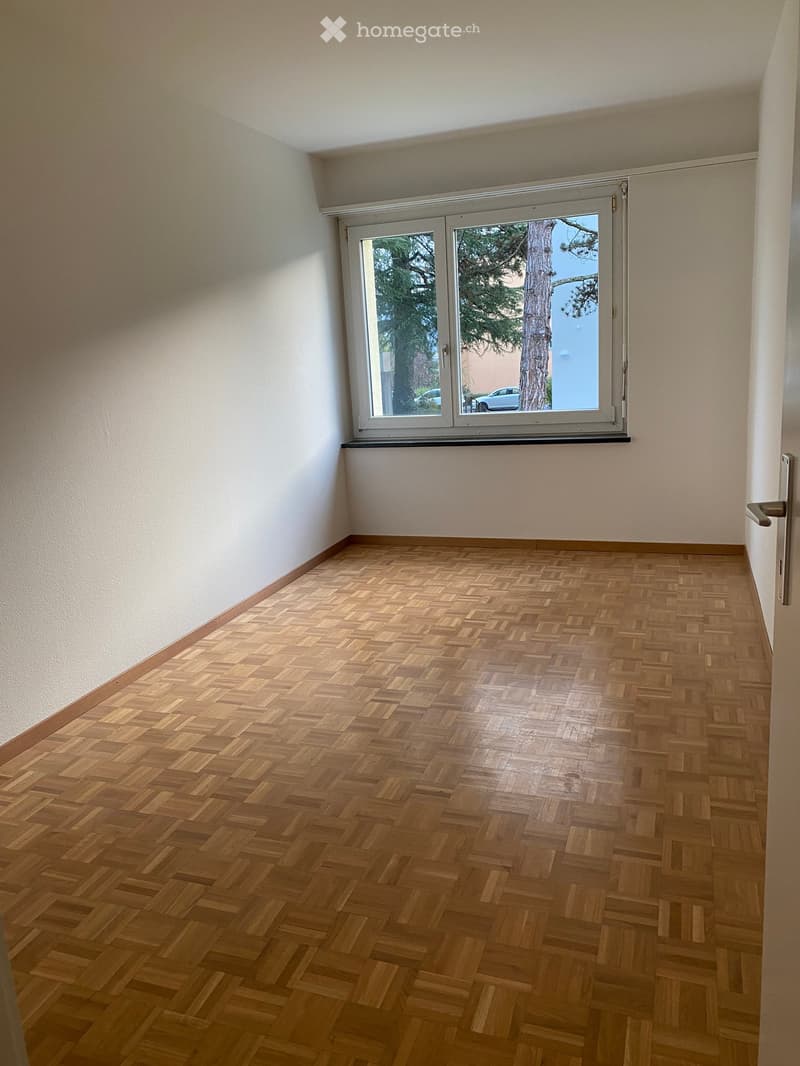 Empty room with wooden herringbone flooring, white walls, and a large window with a view of trees and parked cars.