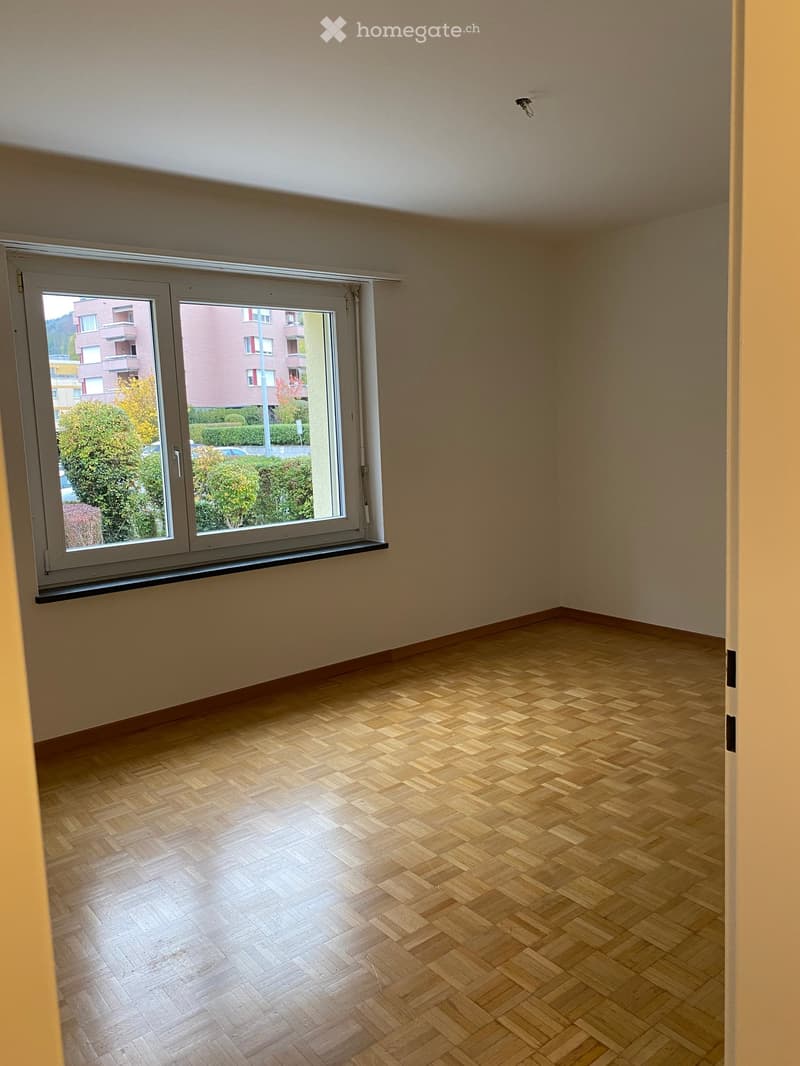 Empty room with wooden herringbone flooring and large window offering view of buildings and greenery outside.