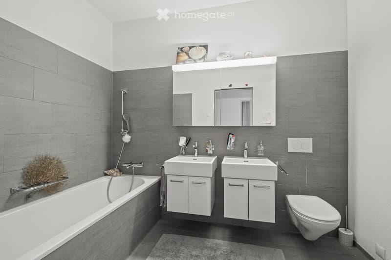 Modern bathroom with grey tiled walls, white fixtures including a bathtub, double sink vanity, toilet, and a large mirror above the sinks. The space features a sleek, contemporary design with a decorative plant in the corner.