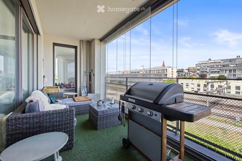 Modern balcony with a view, featuring a wicker sofa, small table, chairs, and a grill.