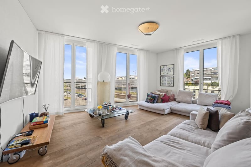 Modern living room with large windows offering a view, white walls, light wooden flooring, a white sectional sofa, a glass coffee table, and decorative items on a side table.