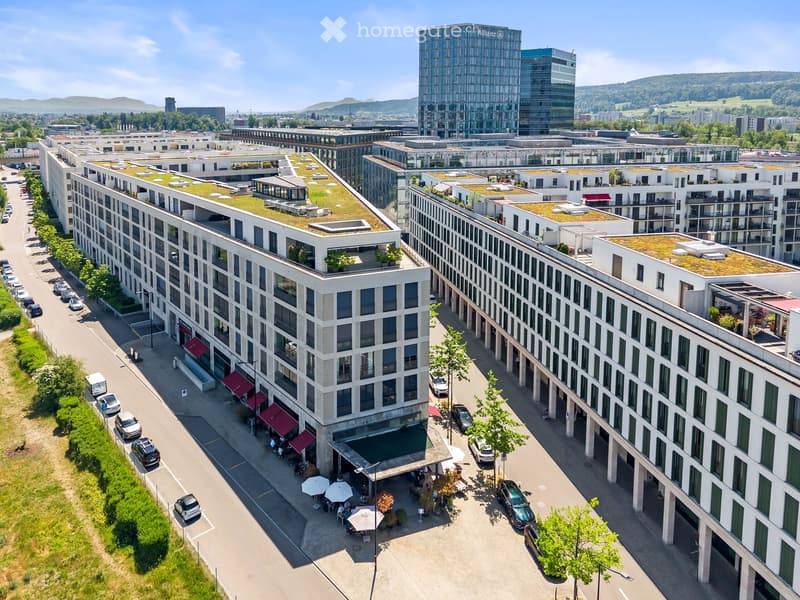Aerial view of a modern urban complex with multiple buildings, green roofs, parking areas, and surrounding greenery.