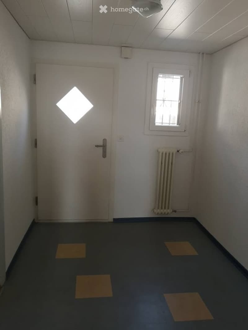 Empty room with white walls, a single window, a white door with a diamond-shaped light fixture, a radiator, and a dark blue carpeted floor with square yellow tiles.