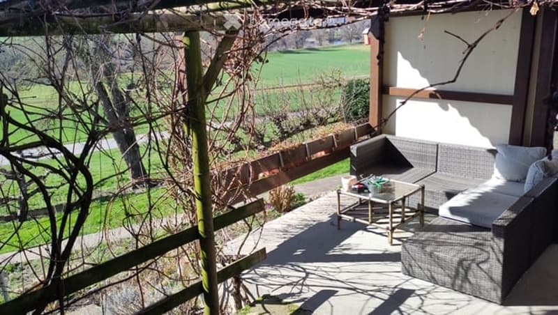 Outdoor patio with a view of a green field, featuring a wooden fence, a small table, and a grey sofa with cushions.