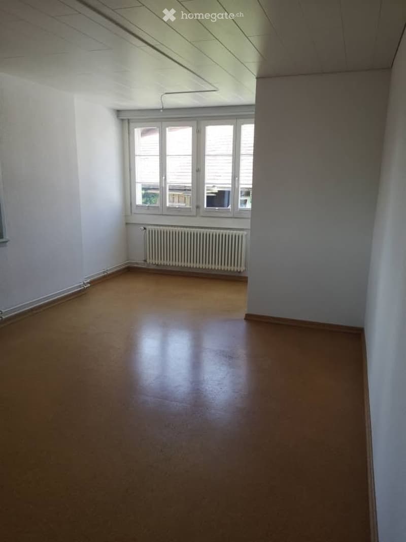 Empty room with wooden flooring, white walls, and a radiator under a large window. No furniture or equipment present.