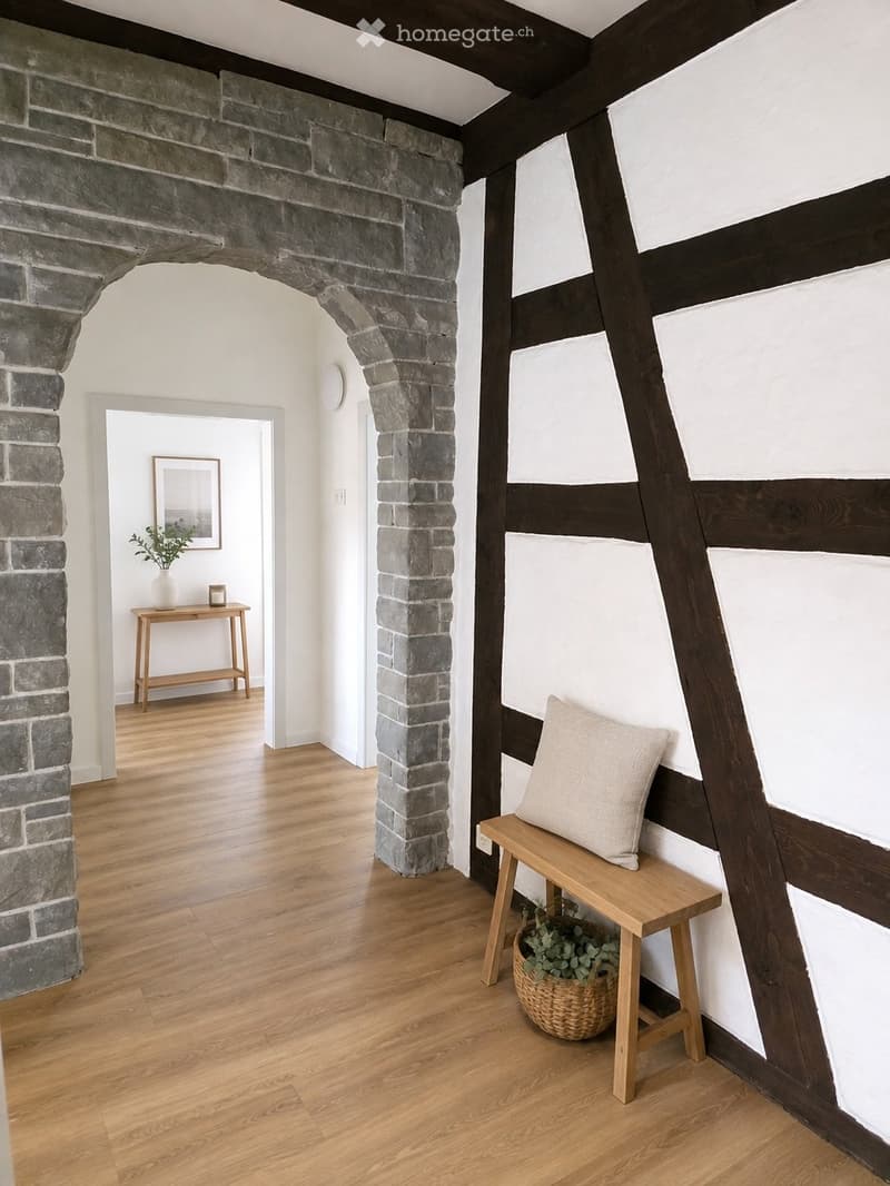 A modern hallway with wooden flooring, exposed wooden beams, and a stone pillar. Features include a small wooden bench with a cushion, a woven basket with plants, and a small wooden table with a vase and candle in the adjacent room.