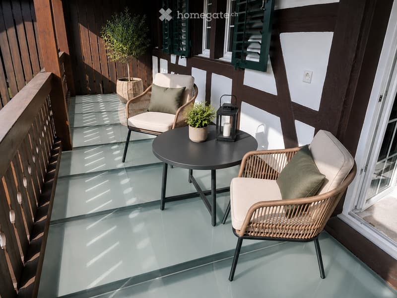 Balcony with two wicker chairs, a round black table, potted plants, and a lantern, featuring a wooden railing and glass floor.