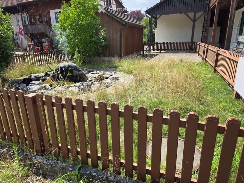A small garden area enclosed by a wooden fence, featuring a stone pathway, a small water fountain, and a mix of grass and rocks.