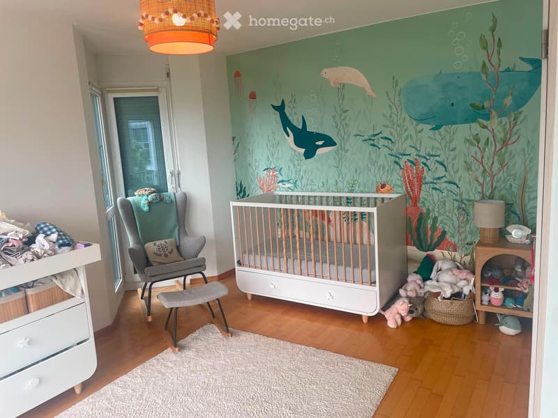 A well-lit children's bedroom with a crib, a rocking chair, a small table, and a shelf filled with toys. The walls feature an ocean-themed mural with fish and seaweed.