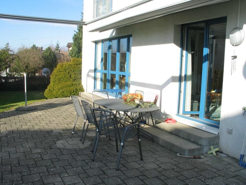Brick-paved patio with a round table, four chairs, and a flower pot, adjacent to a white house with large blue-framed windows and a sliding glass door.