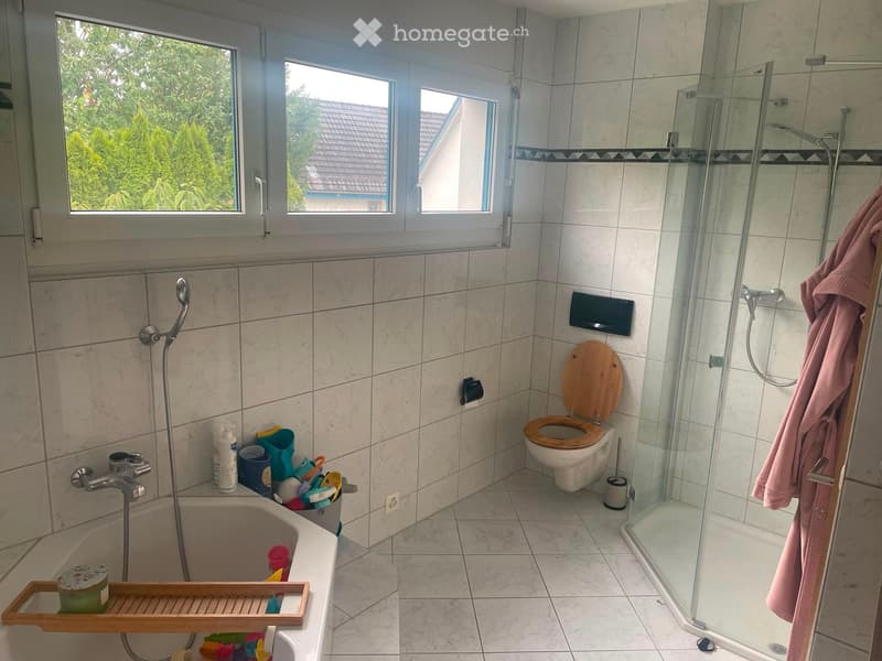 Bathroom with a toilet, bathtub, shower, and various toiletries. The room features tiled walls and a window with a view of greenery outside.