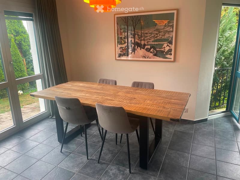 Dining room with a large wooden dining table and four grey chairs, large windows with curtains, and a framed painting on the wall.
