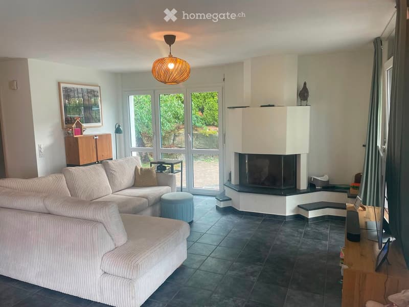 Modern living room with a large sectional sofa, a fireplace, a pendant light, and large windows overlooking a garden.