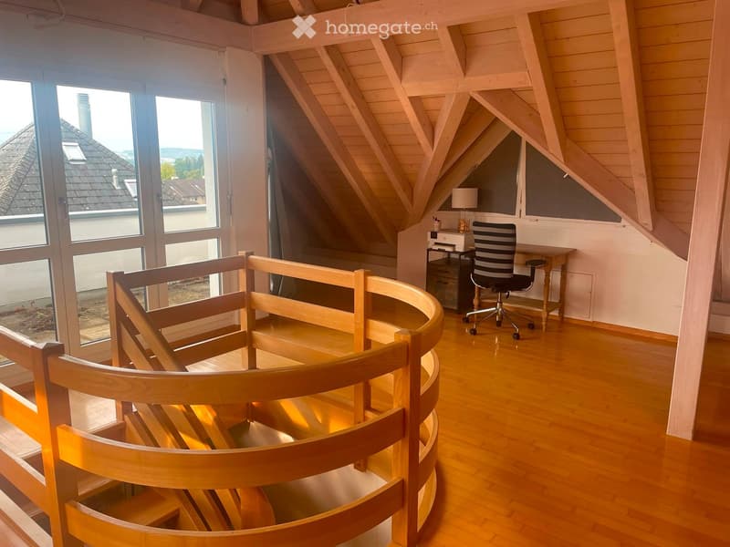 Spacious room with wooden floors, large windows, a curved staircase railing, a desk with a chair, and a lamp on a small table.